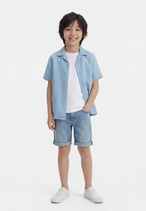 Ένα αγόρι που χαμογελά στέκεται με το ένα χέρι στην τσέπη φορώντας ανοιχτό μπλε πουκάμισο με κοντά μανίκια και κουμπιά, λευκή μπλούζα, τζιν σορτς και λευκά αθλητικά παπούτσια.