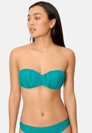 Mujer de cabello largo y oscuro, vestida con un bikini turquesa sin tirantes, con la parte superior e inferior a juego, posando frente a un fondo liso.