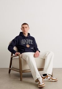 Sudadera navy con el logo "GANT", jeans blancos ajustados y zapatillas marrones y blancas. Modelo sentado en una silla de madera frente a un fondo sencillo.