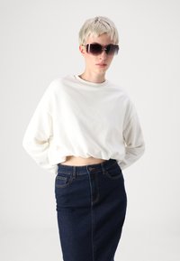 Fehér cropped sweatshirt kerek nyakkal, párosítva egy magas derékú sötét farmer szoknyával. A modellen nagyméretű, sötét napszemüveg van.