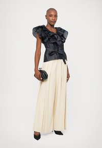 Svart blus med volanger och satinliknande textur, kombinerad med vida, veckade beige byxor. En svart kuvertväska och spetsiga svarta klackar fulländar looken.