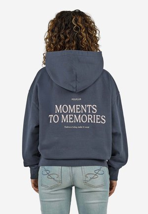 Person med krøllet hår iført en mørkeblå hættetrøje med teksten "MOMENTS TO MEMORIES" på ryggen og lyseblå jeans.