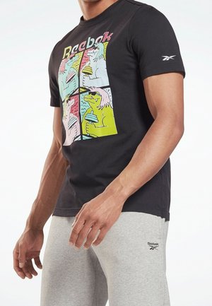 Muž v černém tričku Reebok s barevnými kreslenými psy držícími činky a v šedých teplácích Reebok, stojící s uvolněnýma rukama.