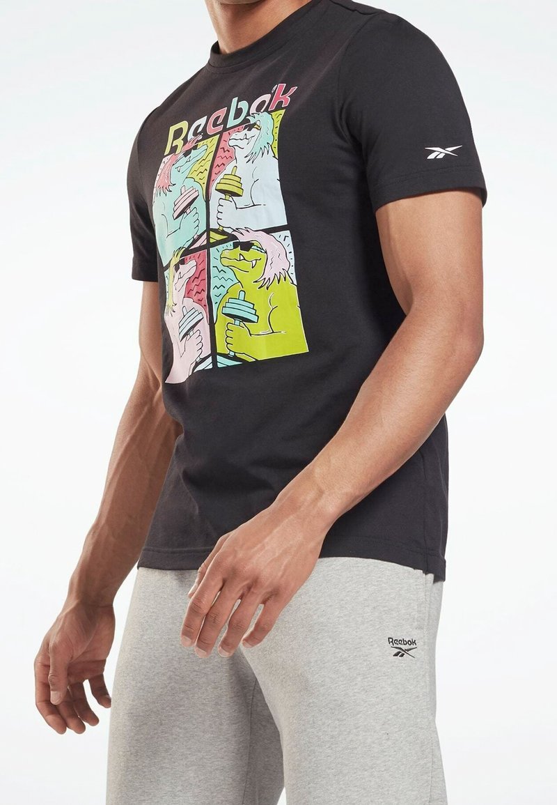 Homme portant un T-shirt noir Reebok avec des chiens de dessin animé colorés tenant des haltères et un pantalon de survêtement gris Reebok, debout les bras détendus.