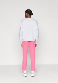 Cilvēks ar muguru, ģērbies gaiši pelēkā sporta jaku, spilgti rozā biksēs, baltās zeķēs un baltās kedās, uz vienkrāsaina fona.