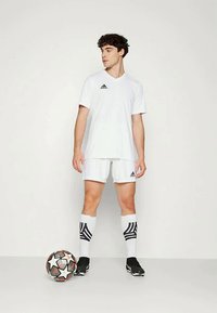 Fehér Adidas foci mez és rövidnadrág, fekete csíkok a zoknin, fekete stoplis, egy színes focilabda mellett áll.