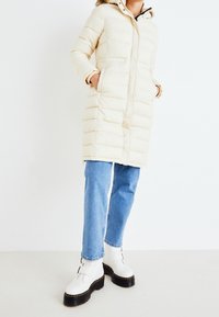 Kremowy puchowy płaszcz z kapturem, z poziomymi przeszyciami, przednimi kieszeniami i zapięciem na zamek, w połączeniu z niebieskimi dżinsami i białymi butami na platformie.