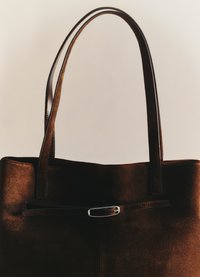 Rudas zomšių pirkinių krepšys su dviem odinėmis rankenomis. Jame yra plokščia forma, elegantiškas dizainas ir sidabro tonų detalė viduryje.