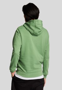 Felpa verde con un fit rilassato, dotata di cappuccio con coulisse, polsini e orlo a coste, e una texture liscia. Indossata sopra una maglietta bianca.
