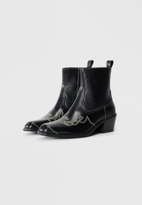Botas de cuero negro hasta el tobillo con punteras afiladas, tacones bajos en bloque y costuras decorativas blancas en un diseño western. Incluyen bucles de tirón.