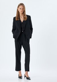 Schwarzer maßgeschneiderter Anzug mit einem strukturierten Blazer, einer taillierten Weste mit Knöpfen und schmalen Hosen, ergänzt durch glänzende schwarze Slingback-Absätze.