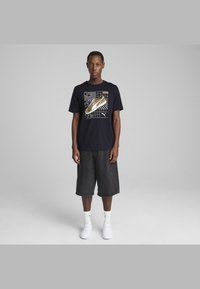 Jūras spēku t-krekls ar grafisku apavu nospiedumu, apvienots ar tumšiem džinsu šortiem. Modelis valkā baltas sneaker un zeķes, demonstrējot ikdienišķu sportisko stilu.
