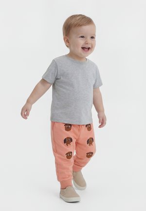 Copil mic zâmbind și mergând, purtând un tricou gri cu mâneci scurte și pantaloni portocalii cu fețe maro de câini, pantofi bej slip-on.