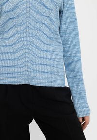 Ljust blå ribbad tröja med strukturerade ränder, med långa ärmar och rundad halsringning, i kombination med svarta byxor.