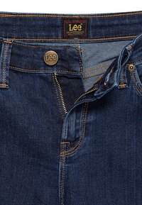 Jean Lee bleu foncé avec fermeture éclair partiellement ouverte, bouton fermé et étiquette de marque visible sur la ceinture.