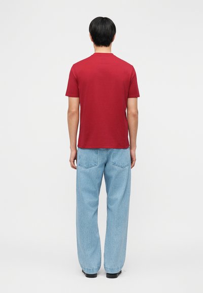 Άντρας, γυρισμένος πλάτη, φοράει απλό κόκκινο T-shirt με κοντά μανίκια, ανοιχτό μπλε τζιν με φαρδιά πόδια και μαύρα παπούτσια, μπροστά σε λευκό φόντο.