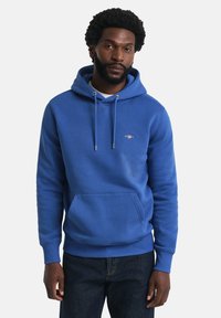 Blå hoodie lavet af en bomuldsblanding, med en frontkangaroo-lomme, snore og ribbede manchetter og kant. Subtilt logo på brystet.
