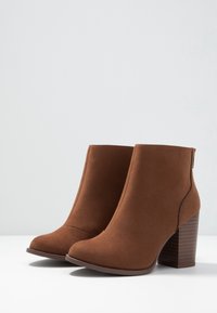 Brązowe zamszowe botki na słupku, z zaokrąglonym noskiem i dekoracyjnym przeszyciem. Obcas ma fakturę przypominającą drewno.