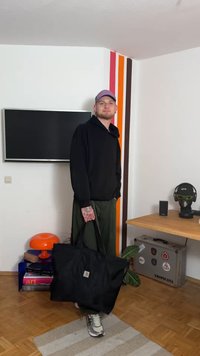 Zwarte stoffen tas met twee handvatten, gedragen door een persoon in een zwart hoodie en groene broek, staand in een kamer met kleurrijke muurbekleding.