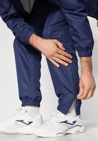 Marineblaue Trainingshosen mit elastischen Bündchen, seitlichen Reißverschlüssen und einer glatten Textur, kombiniert mit weißen Sportschuhen.