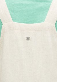 Εκρού λινό ποδιά με φαρδιές τιράντες πάνω από ύφασμα σε μέντα πράσινο, με ένα μικρό στρογγυλό μεταλλικό κουμπί στο κέντρο.