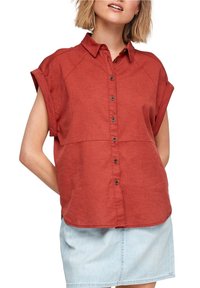 Femme portant une chemise orange rouille à manches courtes avec manches retroussées et une jupe en jean claire, les bras derrière le dos.