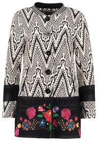 Svartvitt mönstrad lång kappa med en texturerad zickzackdesign, prydd med svarta knappar och färgglad blomsterbroderi vid fållen.