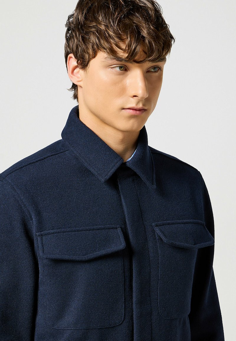 Giacca camicia in lana navy con colletto, due tasche anteriori a patta e una finitura testurizzata. Il design è attillato con una silhouette ben definita.