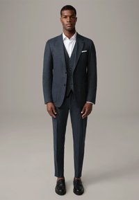 Completo scuro in tessuto a trama fine, con una vestibilità slim, una camicia bianca e mocassini neri. Presenta una giacca a un petto e un gilet a tre bottoni.