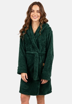 Giovane donna con capelli lunghi e ondulati che sorride, indossando un accappatoio di velluto verde scuro legato in vita su uno sfondo chiaro e uniforme.