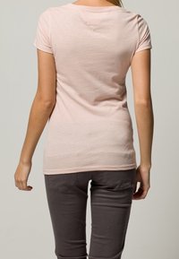 Svetle růžové tričko s krátkým rukávem a kulatým výstřihem, s malým trojúhelníkovým detailem na zadní straně, spojené se šedými kalhotami.