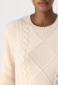 Pull en tricot beige clair avec un col rond côtelé. Présente des motifs en relief de câbles et de diamants sur le devant. Tissu doux.