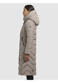Lungo cappotto puffer beige con cappuccio, caratterizzato da un design trapuntato e un motivo a zig-zag. Include tasche con zip e dettaglio del logo sulla manica.