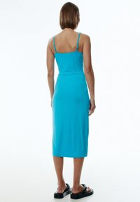 Ein ärmelloses, rückenfreies, knielanges Kleid in leuchtendem Türkis, aus einem glatten Stoff mit dünnen verstellbaren Trägern und einer taillierten Silhouette.