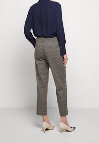 Žena v šedých kostičkovaných kalhotách, námořnické dlouhé blůze a béžových nízkých podpatcích, stojící na neutrálním pozadí.