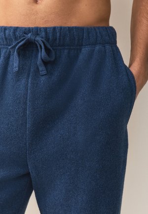 Muž nosící tmavě modré fleecové tepláky s elastickým pasem a stahovací šňůrkou, ruka v kapse, nahý trup viditelný nad pasem.