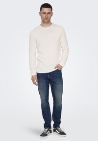 Weißes langärmliges Textshirt, blaue Jeans und dunkle Sneakers mit weißen Akzenten. Modell steht vor einem hellen Hintergrund.