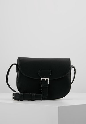 Sac bandoulière en cuir noir avec rabat et fermeture à boucle argentée, bandoulière ajustable, exposé sur un piédestal blanc devant un fond gris.