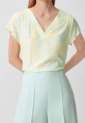 Kvinde iført pastelgul V-hals top med subtilt løvmønster og lyseblå bukser med høj talje, hænderne hvilende på hofterne.