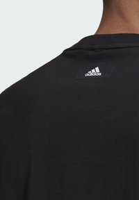 Černé tričko Adidas, na zádech malé bílé logo Adidas blízko výstřihu na horní části zad osoby.