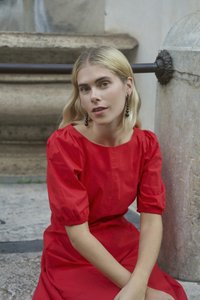 Crvena pamučna haljina s kratkim nabranim rukavima, okruglim izrezom i ravnim krojem. Model ima dugu, valovitu plavu kosu i crne naušnice.