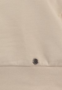 Tessuto beige con una superficie liscia, caratterizzato da un distintivo etichetta argentata con la scritta "Kaye Largo" vicino alla parte inferiore. Design semplice e minimalista.