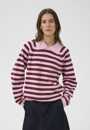 Mujer con un suéter de rayas rosas y burdeos con un cuello ancho y una falda de mezclilla oscura, de pie con las manos en los bolsillos.