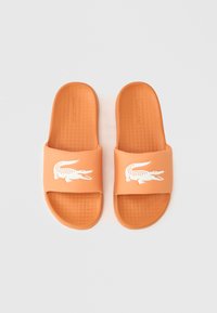 Πορτοκαλί λαστιχένια σανδάλια με υφασμένο πάτο, με λευκό λογότυπο κροκόδειλου στο λουράκι, σχεδιασμένα για καθημερινή χρήση.