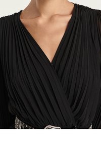 Schwarzes, plissiertes Wickelkleid mit V-Ausschnitt und langen Ärmeln, tailliert mit einem schwarzen, genagelten Gürtel mit silberner Schnalle.
