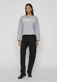 Sudadera gris recortada con el estampado "BROOKLYN" en blanco, combinada con pantalones negros de pierna recta y zapatos negros. Textura suave, corte relajado.