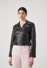 Giubbotto in pelle nera cropped da motociclista con zip e bottoni in argento, caratterizzato da un colletto strutturato e un design aderente, abbinato a pantaloni rosa chiaro.