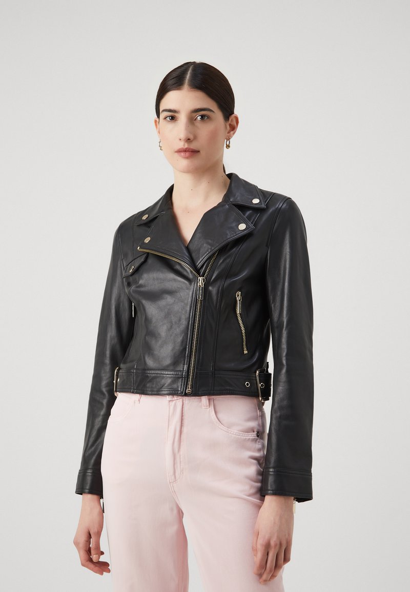 Giubbotto in pelle nera cropped da motociclista con zip e bottoni in argento, caratterizzato da un colletto strutturato e un design aderente, abbinato a pantaloni rosa chiaro.