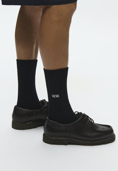 Jambes portant des chaussettes côtelées noires avec le logo « W.W. » et des chaussures en cuir noires à lacets sur un fond clair.