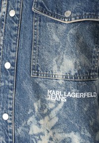 Farkkutakki vaaleansinisen pesun kanssa, jossa on rintatasku ja valkoista brodeerattua tekstiä "KARL LAGERFELD JEANS". Hopeiset napit ovat näkyvissä.